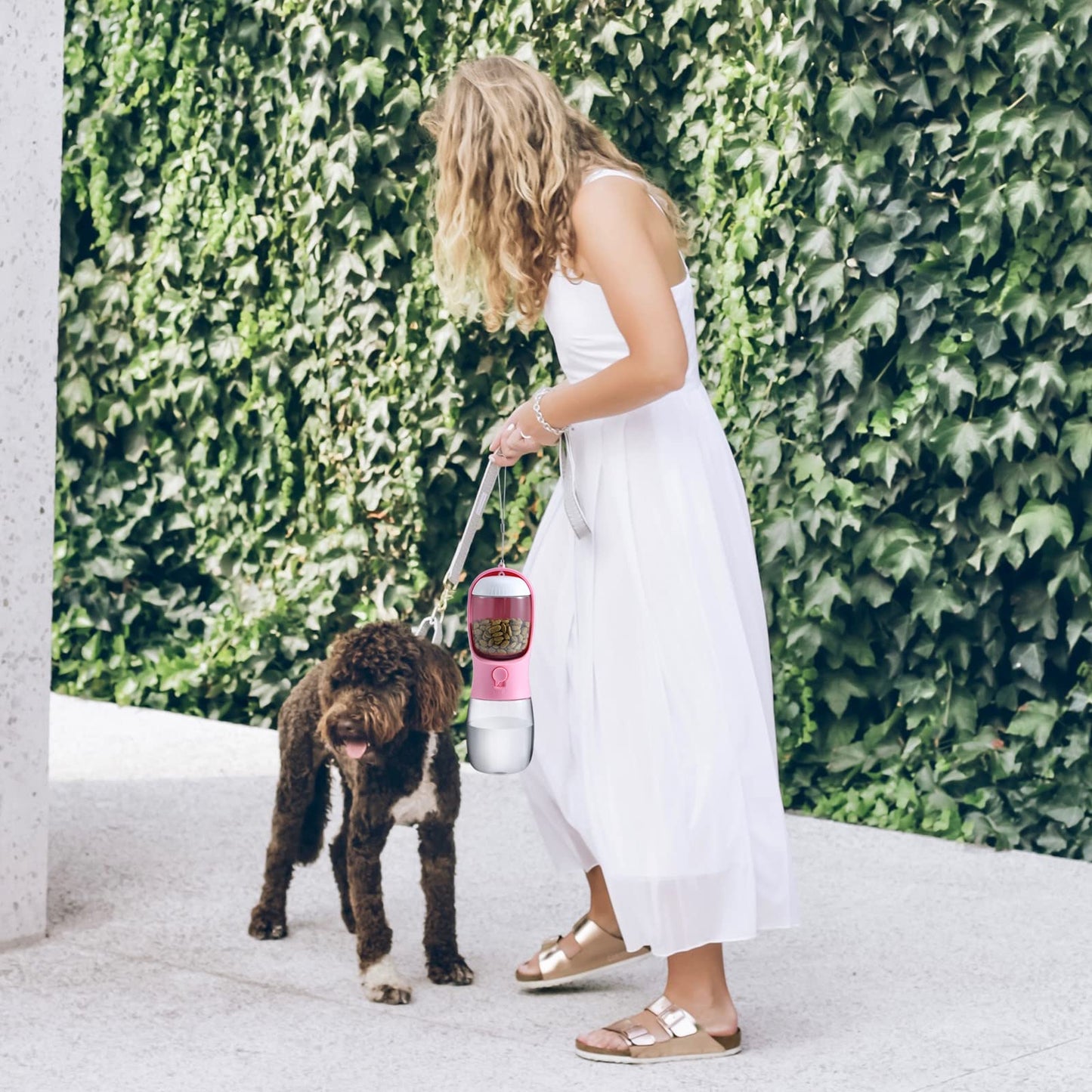 Woman in a white dress holding a pink pet water bottle with a dog standing next to her against a green hedge.
